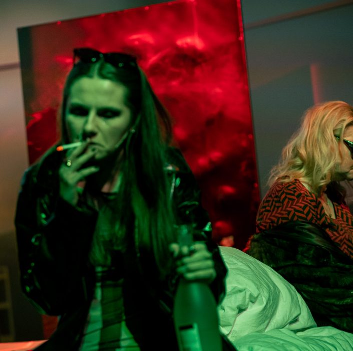 Scena ze spektaklu, kobieta paląca papierosa trzyma w ręku butelkę