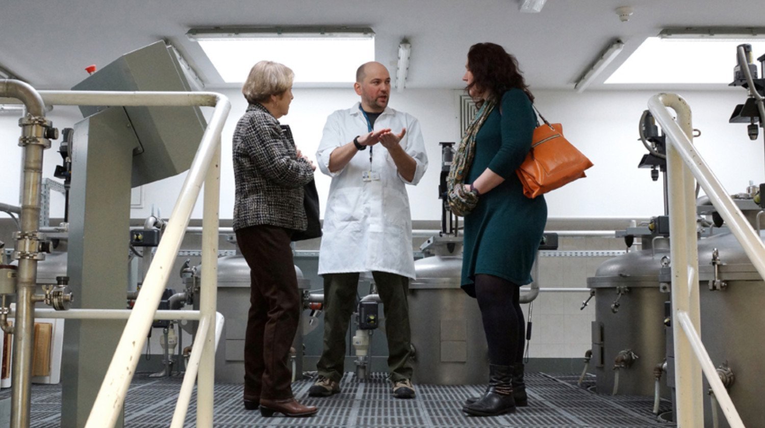 Trójka ludzi rozmawia w laboratorium. Mężczyzna w białym fartuchu tłumaczy coś dwóm kobietom, wskazując rękami.