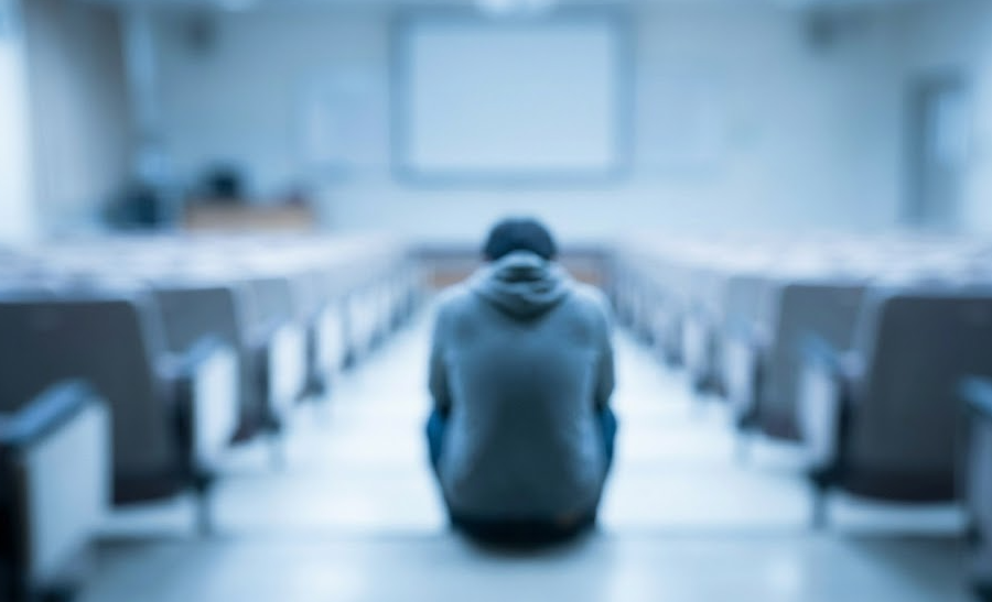 Mocno zamazane zdjęcie w niebieskiej tonacji, przedstawiające samotną osobę siedzącą tyłem na schodach w pustej, dużej sali wykładowej.